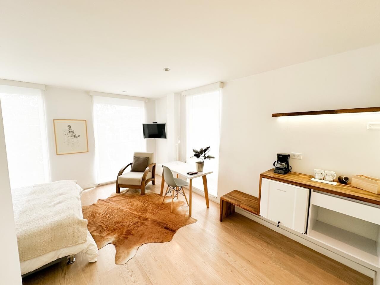 A modern hotel room with a bed, a desk, a chair, a small kitchenette, and a large window.