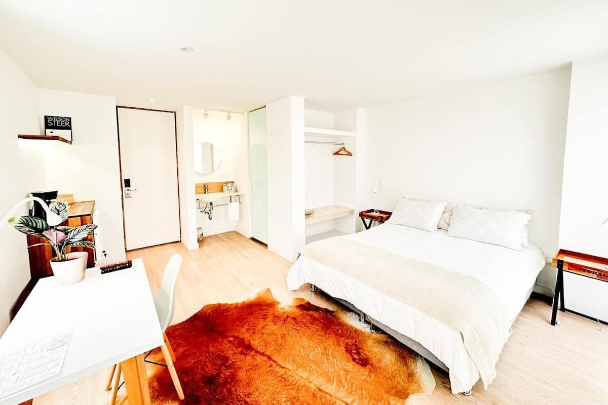 Een moderne slaapkamer met een groot bed, een bureau met stoel, een klein tafeltje en een kapstok.