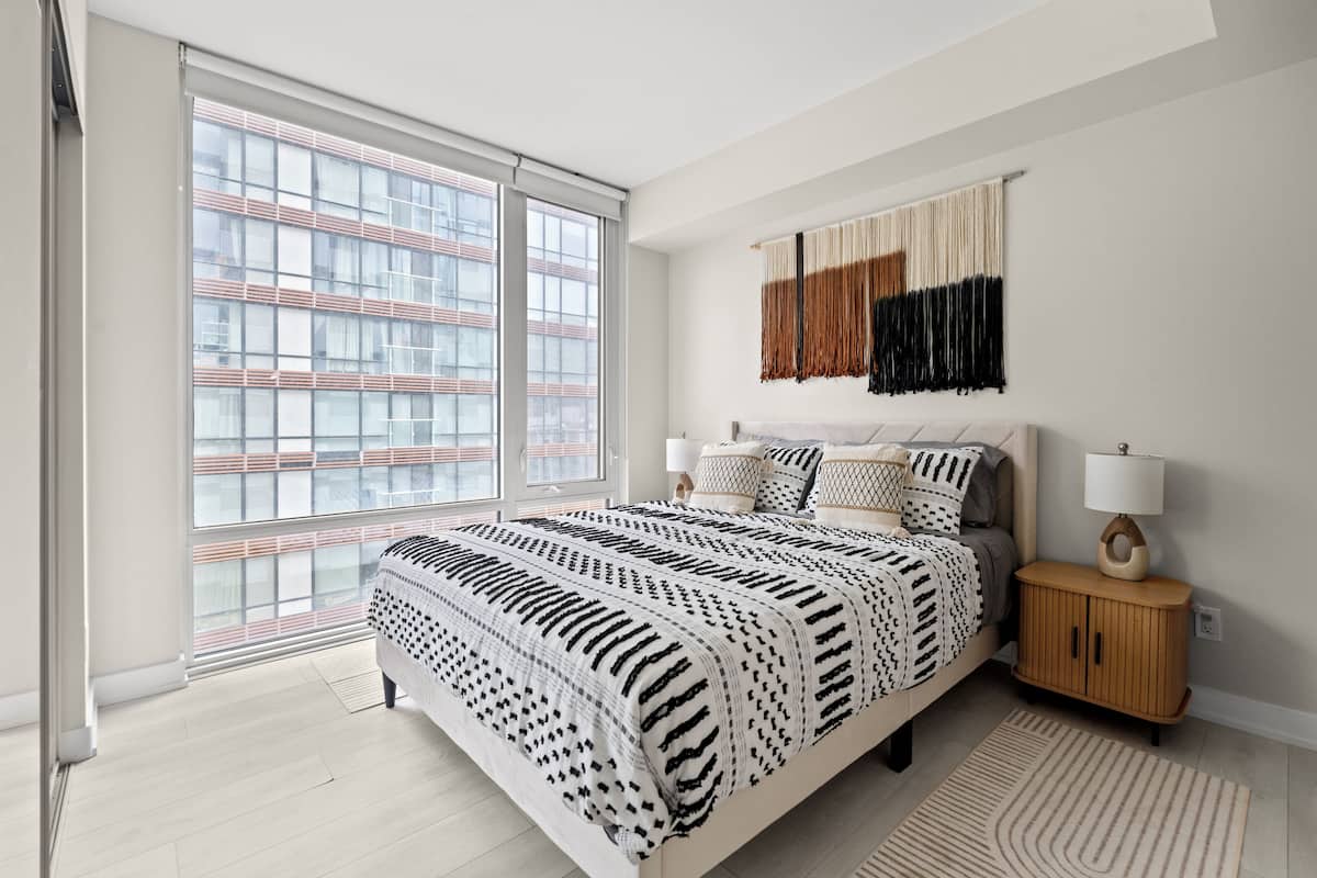 Un dormitorio moderno con un ventanal grande, una cama con ropa de cama estampada, una mesita de noche con una lámpara y una mesita auxiliar de madera.