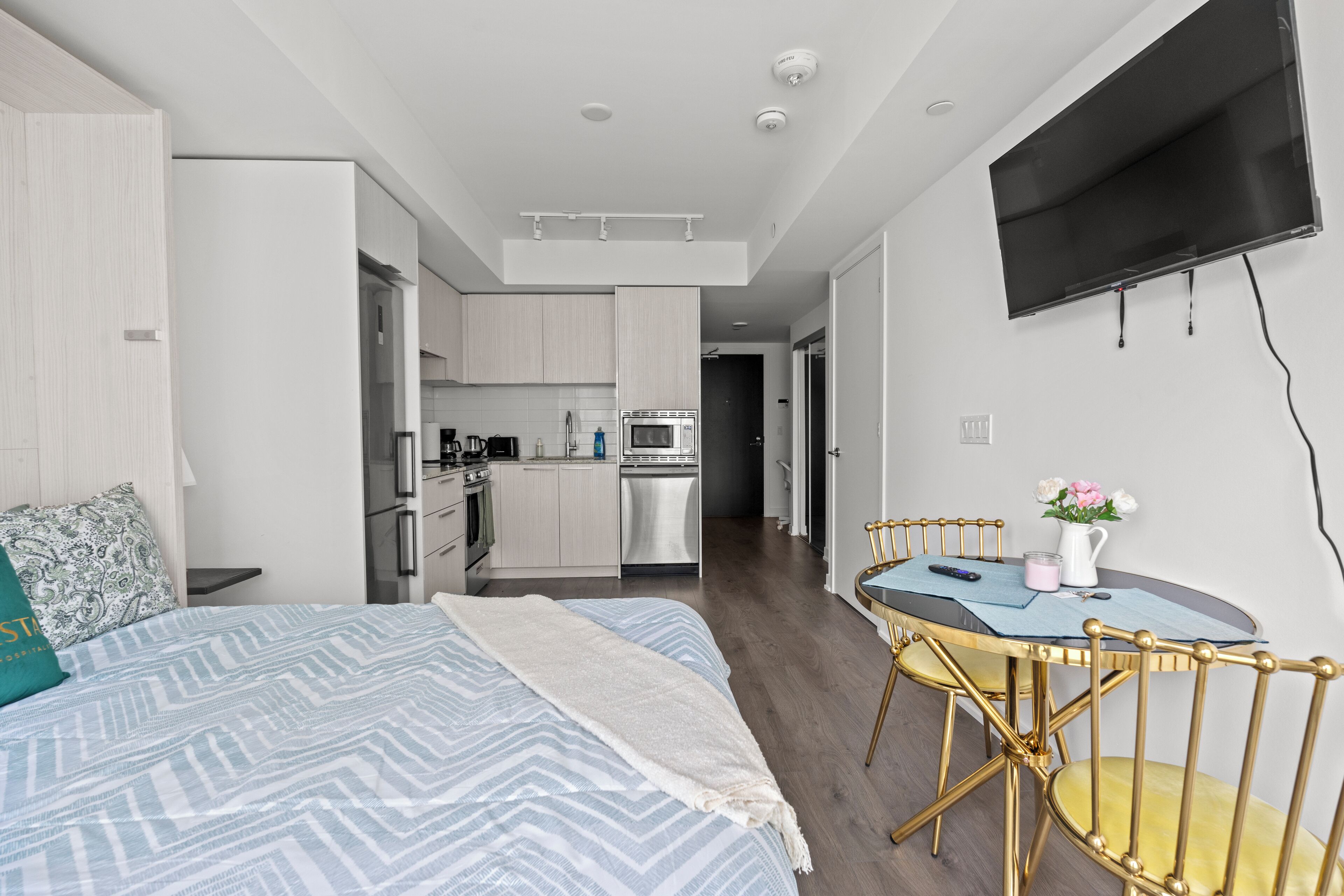 A modern hotel room with a bed, a dining table with chairs, a kitchen area, and a TV mounted on the wall.