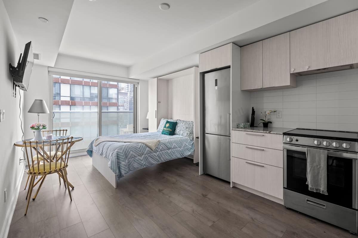 Un dormitorio moderno con cama, mesita de noche, lámpara y vistas a la ciudad a través de un amplio ventanal.