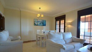 Un salon spacieux avec un ensemble de canapés blancs, une table à manger avec des chaises, un tableau accroché au mur et une grande fenêtre donnant sur un beau paysage.