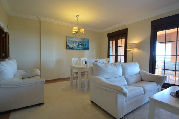 A spacious living room with a white sofa set, a dining table with chairs, a painting on the wall, and a large window with a view.