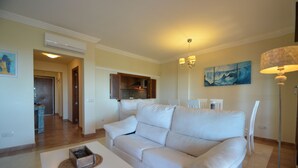 Un salon avec un canapé blanc, une table basse et une lampe. On aperçoit un coin cuisine au fond.