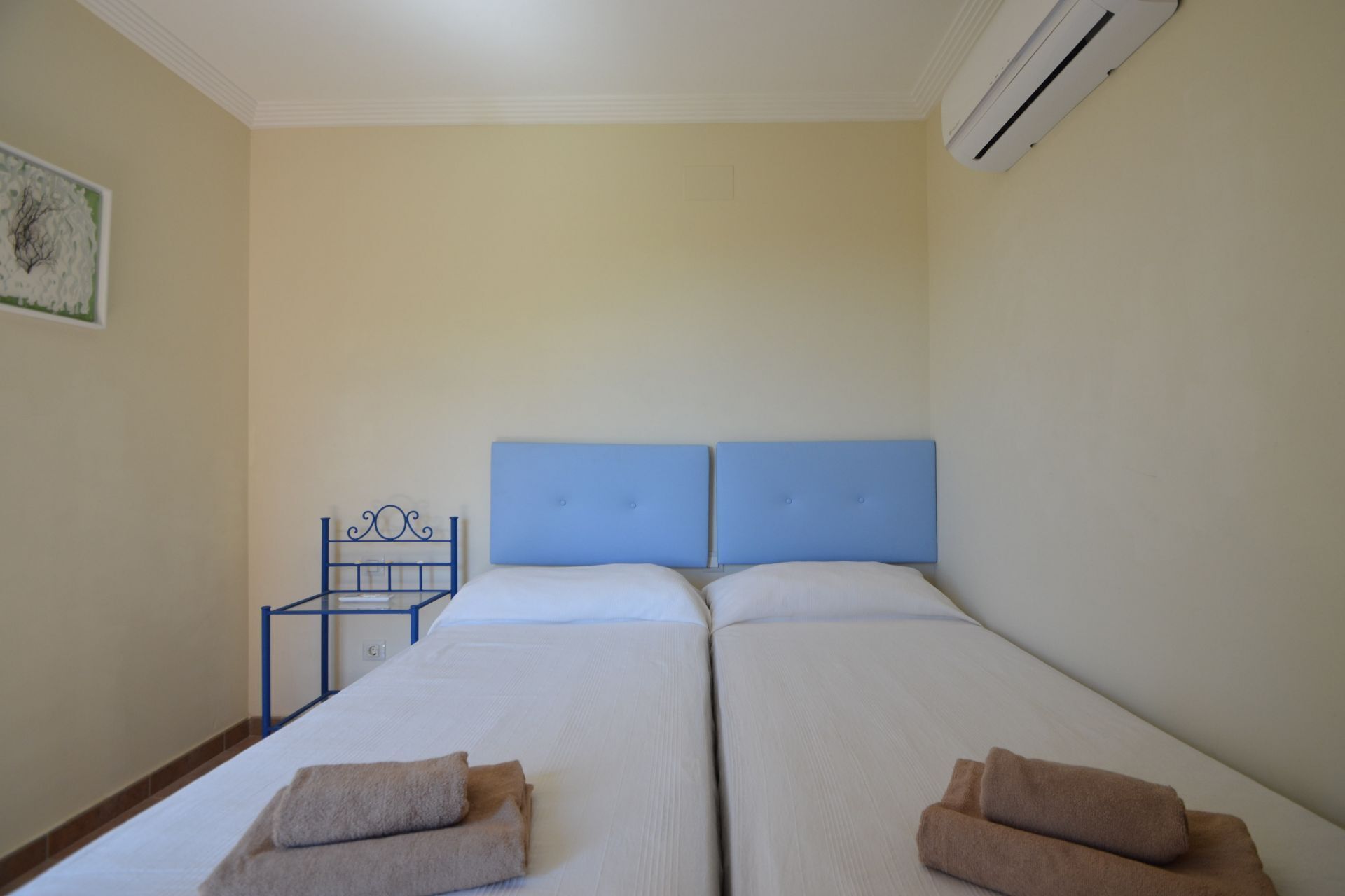 A double bed with a blue headboard, two towels, a small table, and an air conditioning unit.