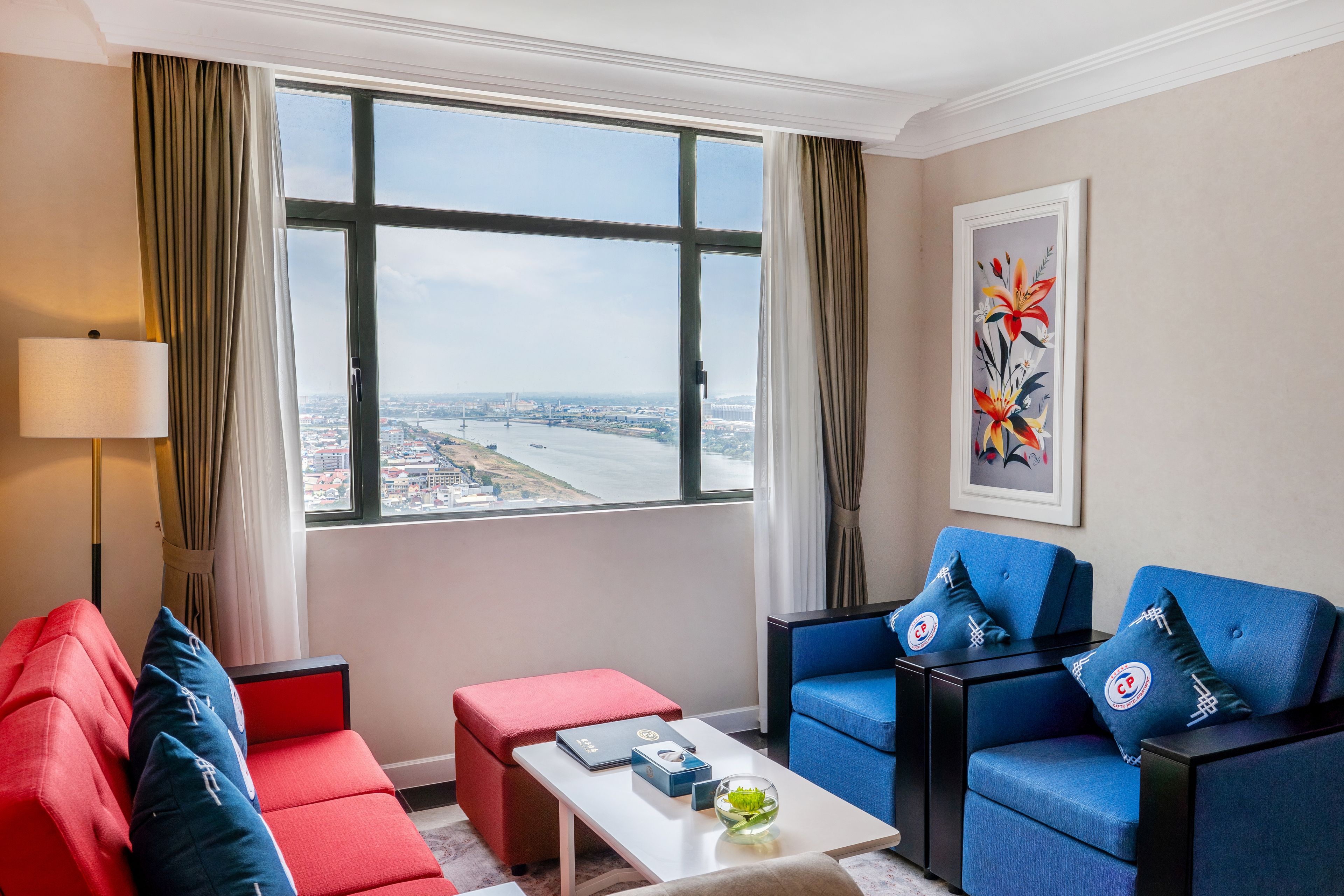 Habitación de hotel con un ventanal grande, un sofá rojo, un sofá azul, una mesita y una lámpara.