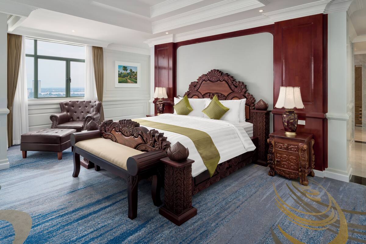 A spacious bedroom with a large bed, a leather armchair, a wooden nightstand, and a view of the city through a large window.