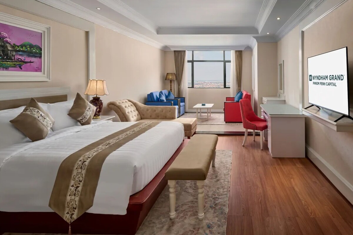 Een hotelkamer met een groot bed, een bureau, een stoel, een klein tafeltje en een televisiescherm waarop de naam van het hotel staat.