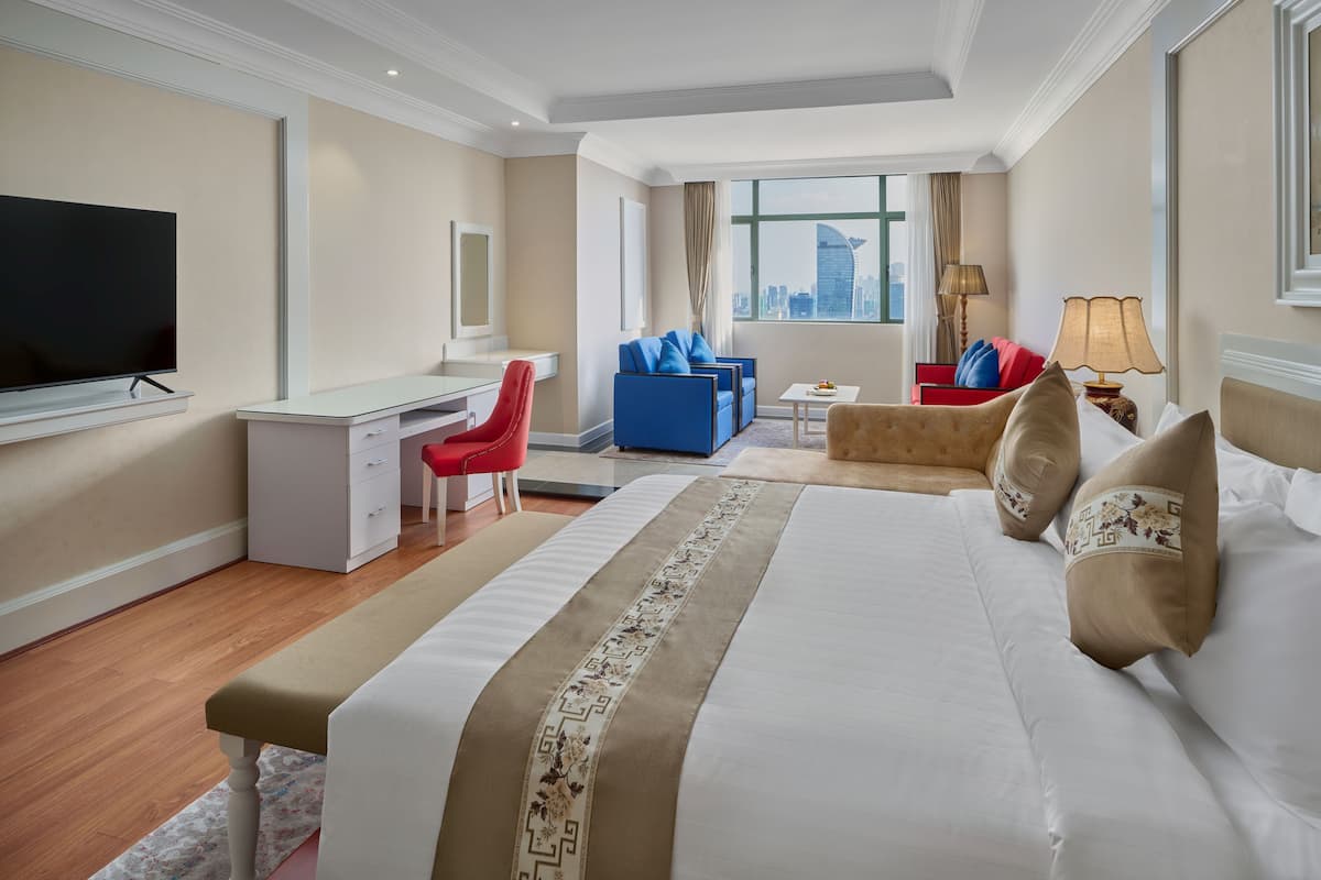 A hotel room with a large bed, a desk, a red chair, a blue sofa, and a view of the city.