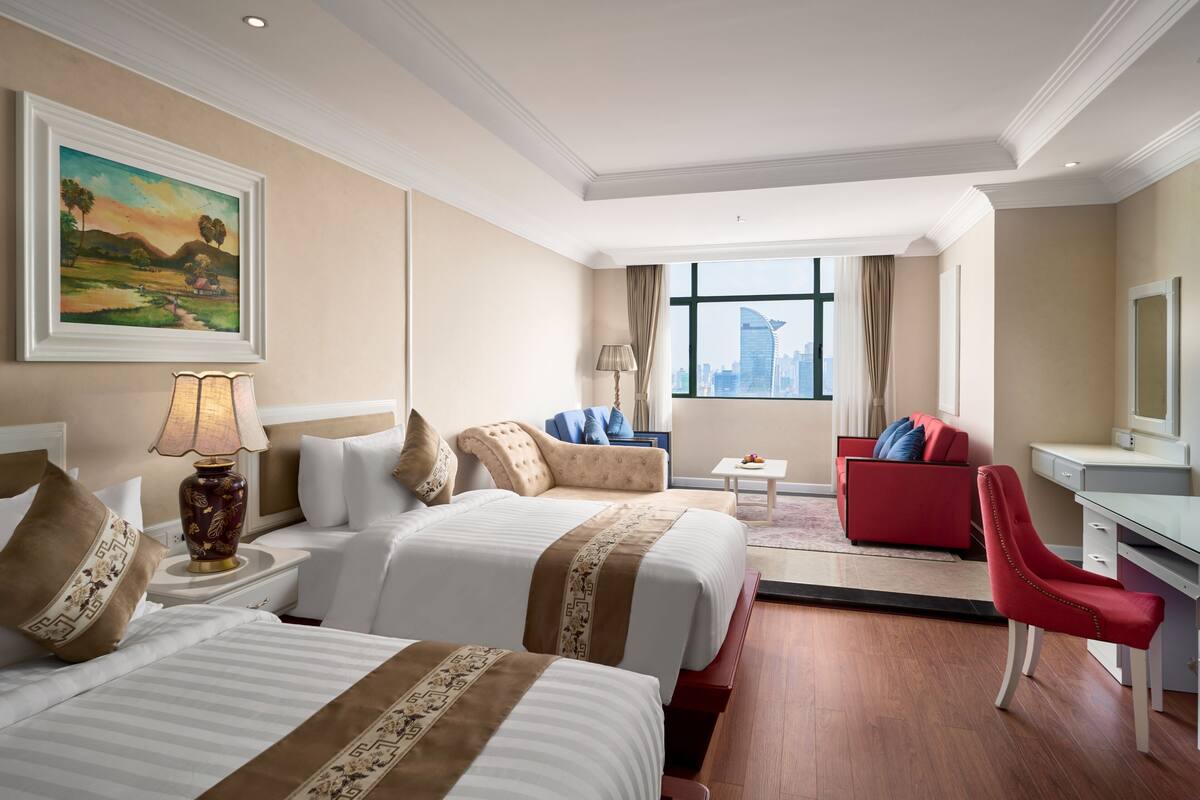 A hotel room with two beds, a red armchair, a desk, and a large window with a city view.