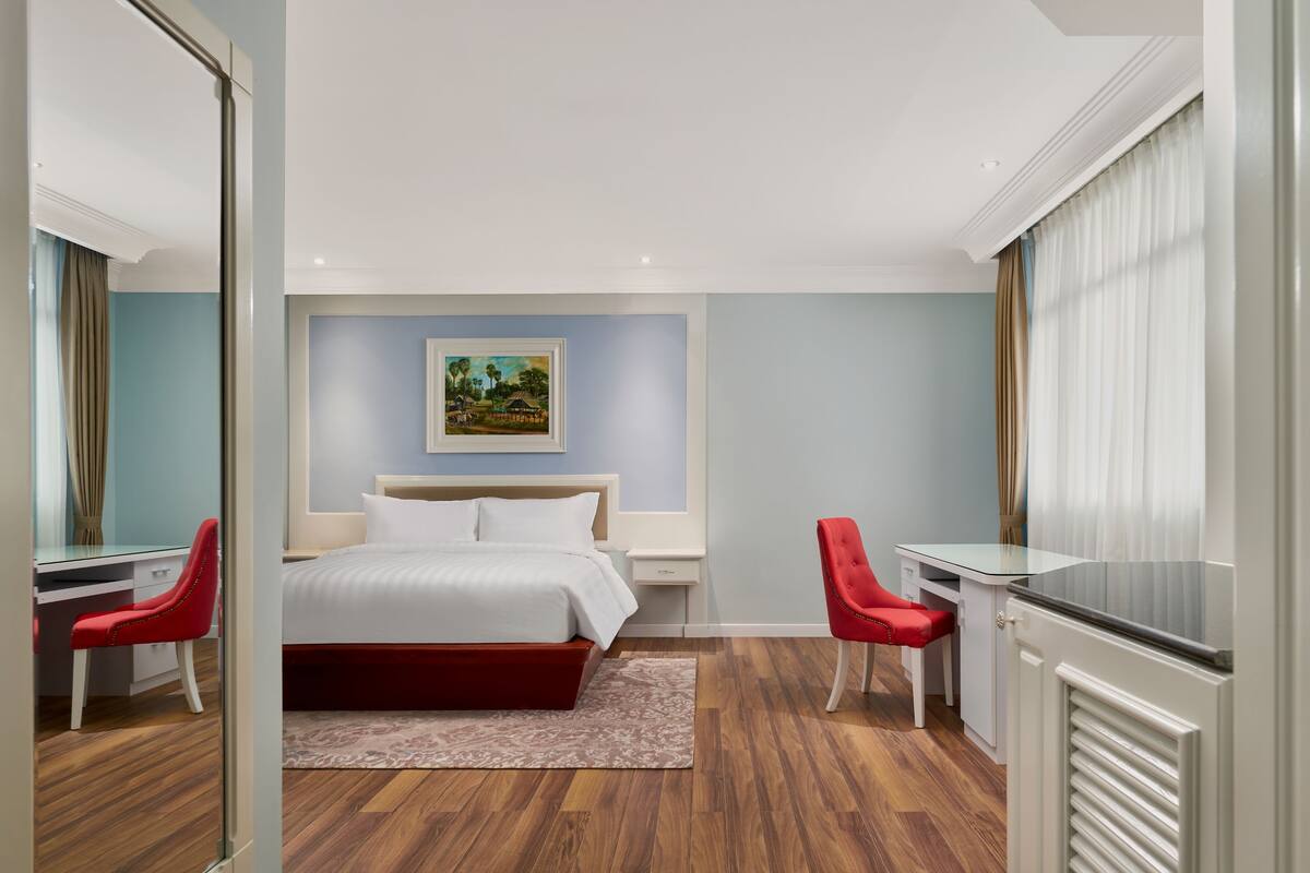 A modern hotel room with a large bed, a desk, two red chairs, and a painting on the wall.