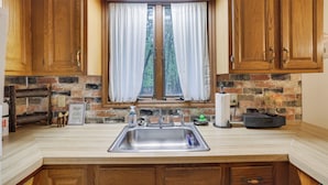 A kitchen with wooden cabinets, a stainless steel sink, a window with white curtains, and a stone backsplash.