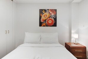 A hotel room with a bed, white walls, a painting of flowers, a nightstand with a lamp, and a wooden drawer unit.