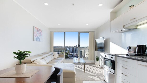 A modern apartment with a dining area, a sofa, a kitchen, and a large window offering a city view. - City Vista Apt with Views, Pool and Gym (Auckland CBD)