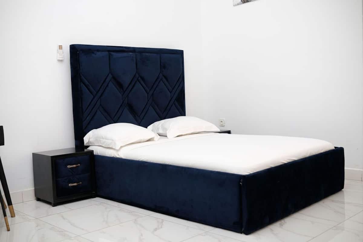 A bed with a dark headboard and white bedding, a bedside table with drawers, and a white wall.