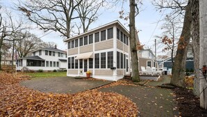 A two-story building with a large windowed front section, a covered porch, and a paved driveway with fallen leaves. - Beautiful, modern cottage with lake views inside Lakeside! (Lakeside)