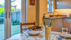 Un coin repas avec une table dressée pour deux personnes, un vase contenant des plantes vertes et, au fond, une cuisine.