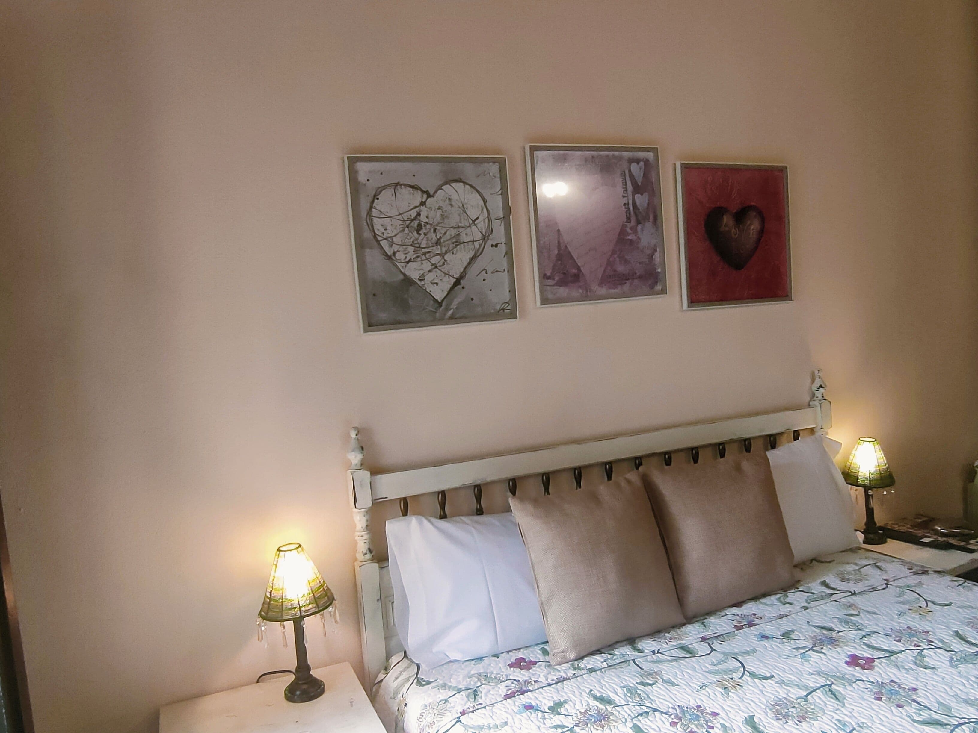 Un dormitori amb un llit, dues làmpades de nit i tres quadres murals amb cor.