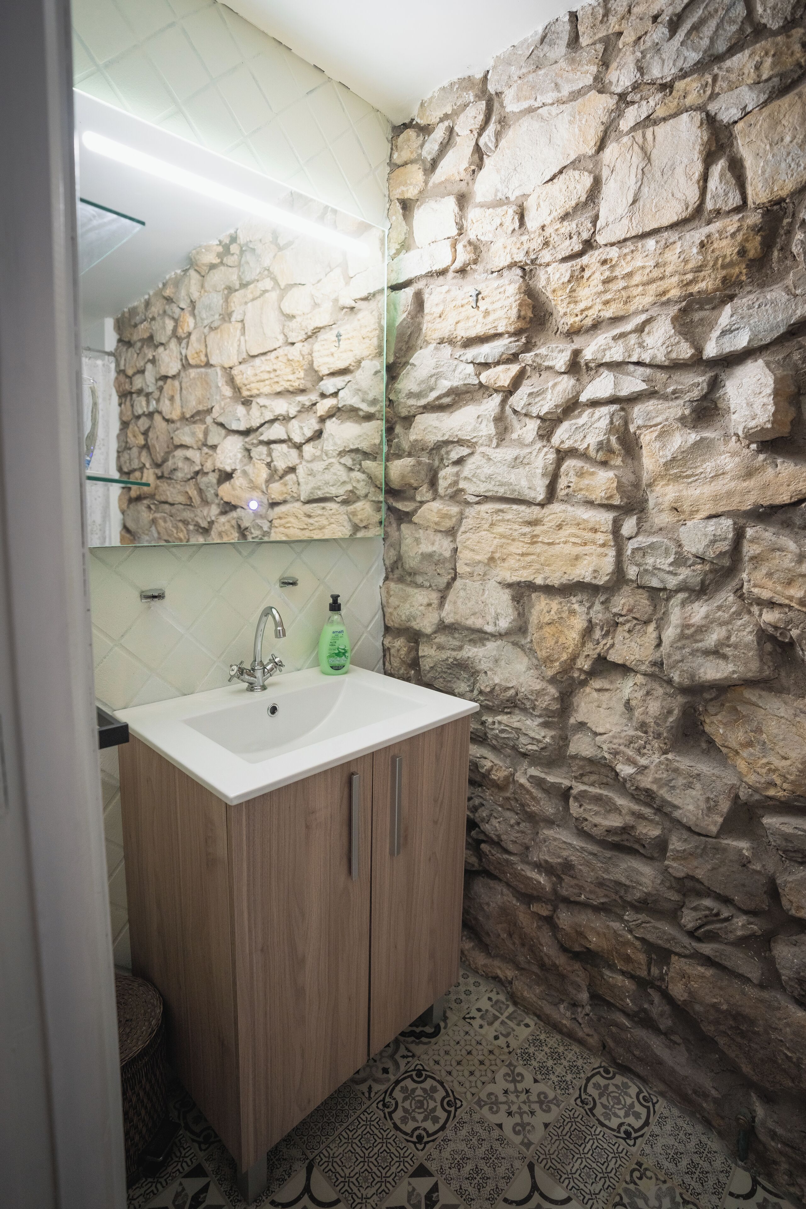 Un baño con pared de piedra, un mueble de madera, un lavabo blanco y baldosas de suelo estampadas.