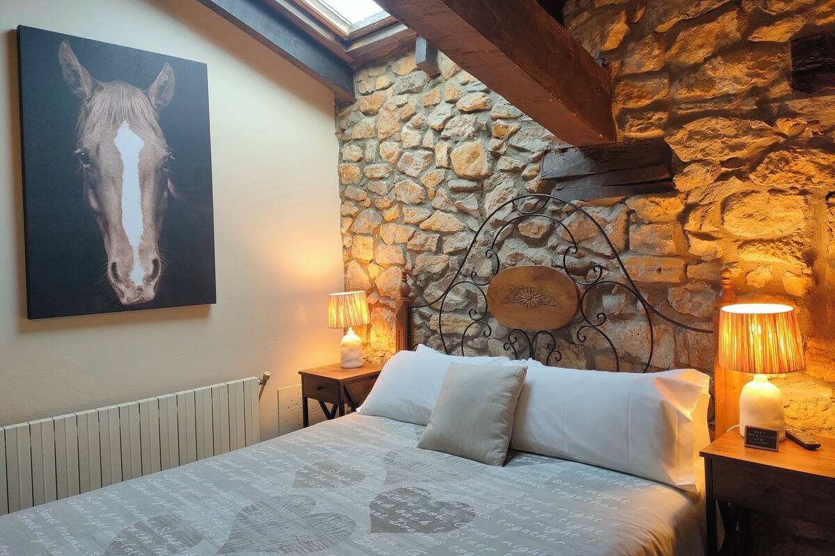 Ein Schlafzimmer mit Steinstruktur, einem Gemälde von einem Pferd, einem Bett mit gemustertem Überwurf, zwei Nachttischlampen und einem hölzernen Kopfteil.