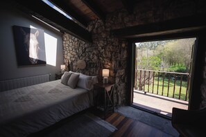 A bedroom with a stone wall, a bed, a nightstand with a lamp, and a view of a garden through a sliding door. - HOTEL RURAL LA JOYUCA DEL PAS (Miengo)