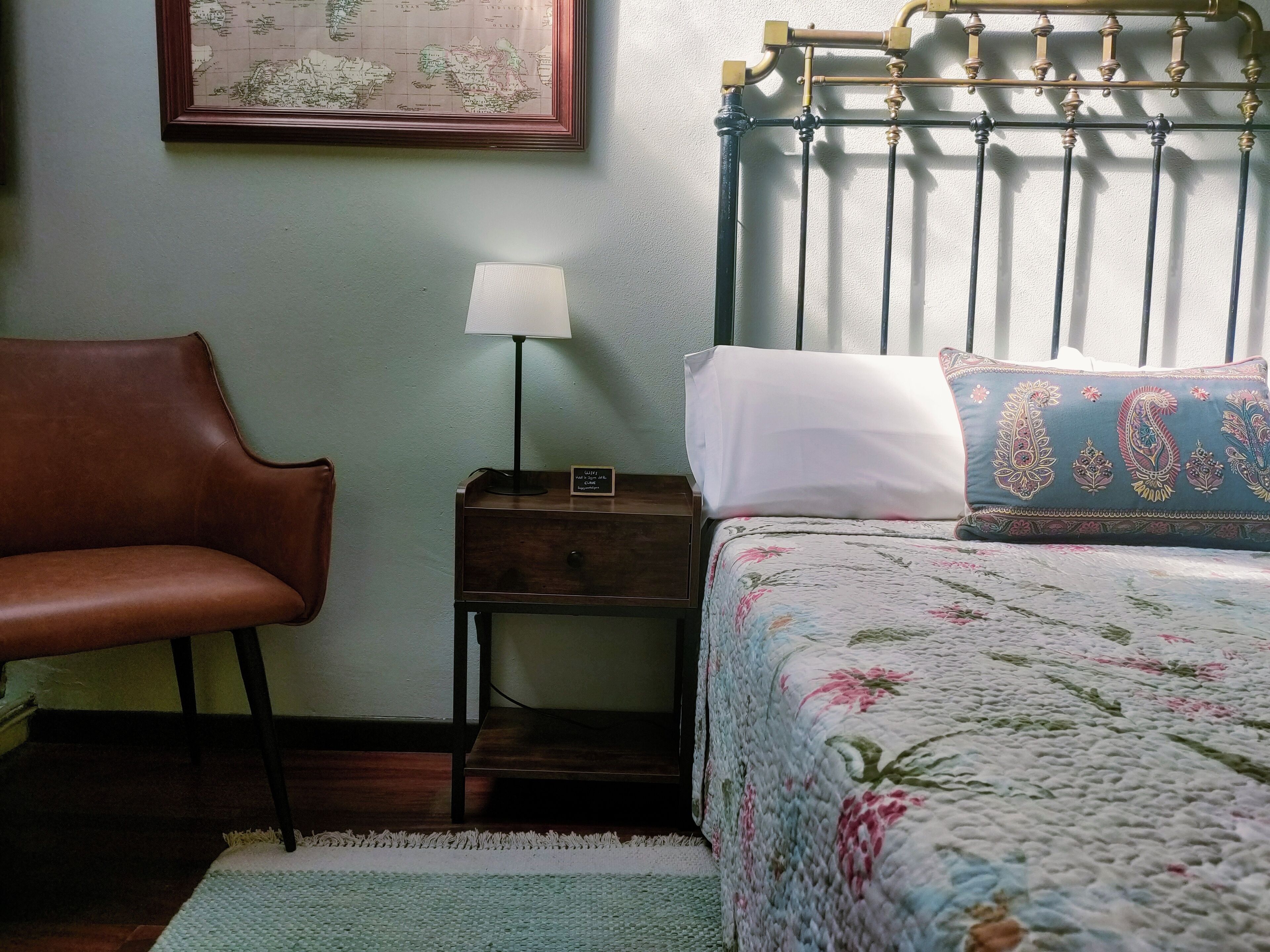 Un dormitorio con cama, mesita de noche, lámpara, silla y un mapa enmarcado en la pared.