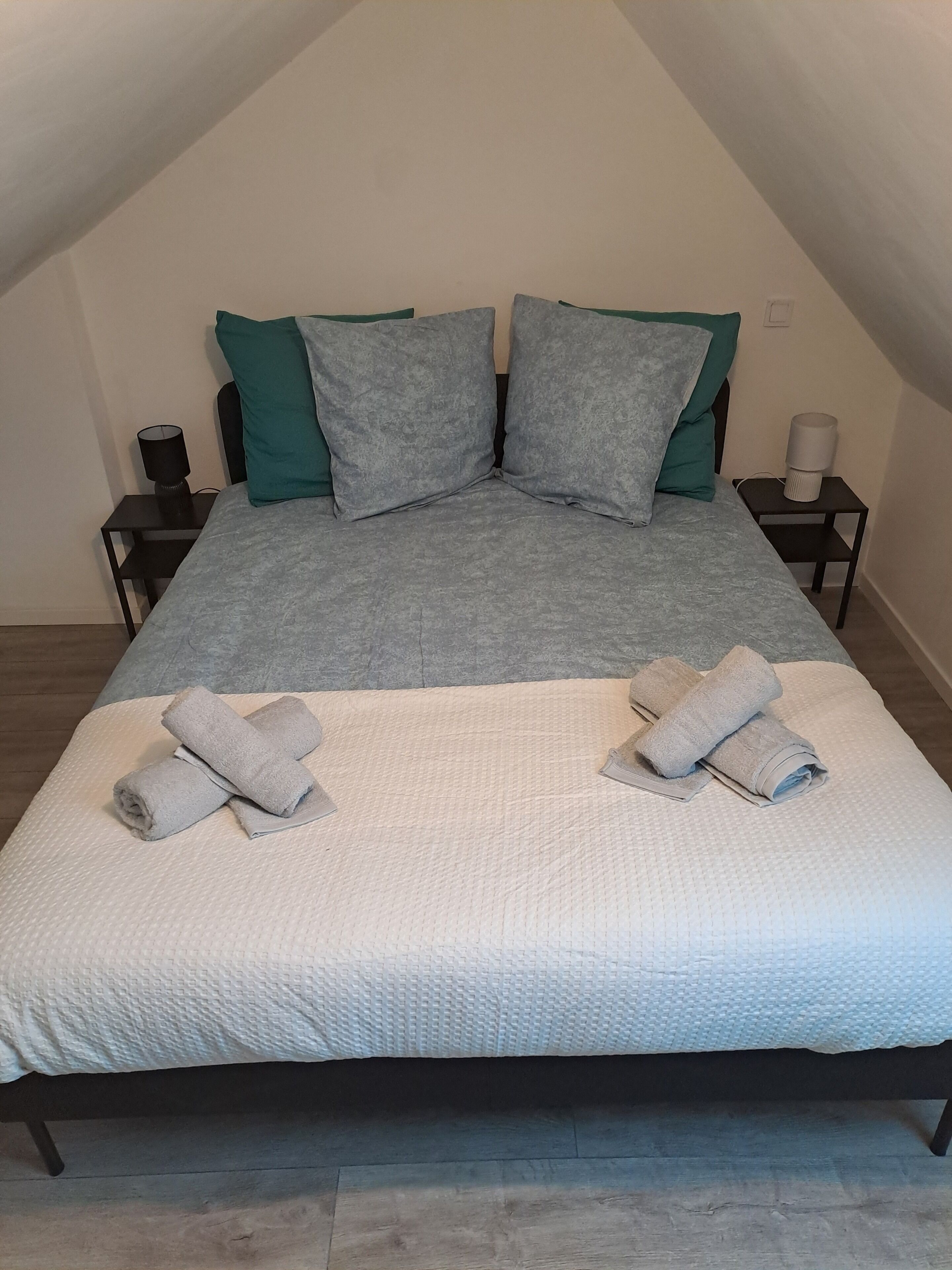 Cama bem arrumada com roupa de cama cinza e branca, dois travesseiros e duas toalhas enroladas em um chão de madeira.