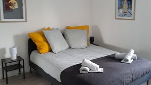 Ein modernes Schlafzimmer mit schwarzem Bettgestell, weißen und gelben Kissen, einer grauen Bettdecke und zwei zusammengerollten Handtüchern auf dem Bett. Ein kleiner Nachttisch mit Lampe und ein gerahmtes Bild an der Wand sind ebenfalls vorhanden.