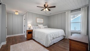 Une chambre à coucher comprenant un lit, des tables de chevet, un ventilateur de plafond, une fenêtre avec des rideaux et une commode en bois.