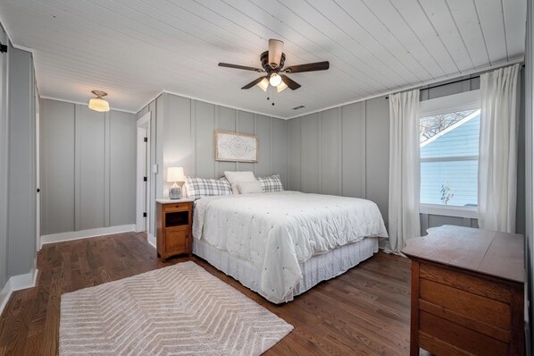 Un dormitorio con cama, mesitas de noche, ventilador de techo, ventana con cortinas y cómoda de madera.