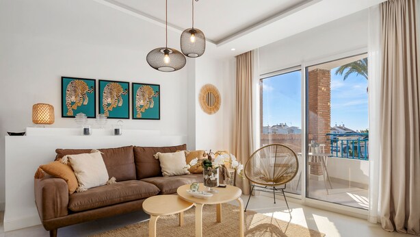 A modern living room with a brown sofa, a round wooden coffee table, and a wicker chair. There are three wall-mounted pendant lights, a large window with a view of buildings, and a balcony with a chair. - Colorful Flat in Riviera del Sol Ref 27 (Mijas)