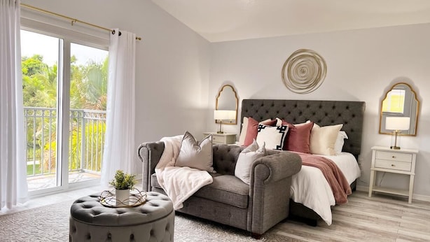 Un dormitorio con un sofá gris, un otoman tapizado, una cama con varias almohadas, dos mesitas de noche con lámparas, y un ventanal con cortinas.