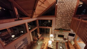 A spacious living area with a stone fireplace, wooden furniture, and large windows offering views of the outdoors.