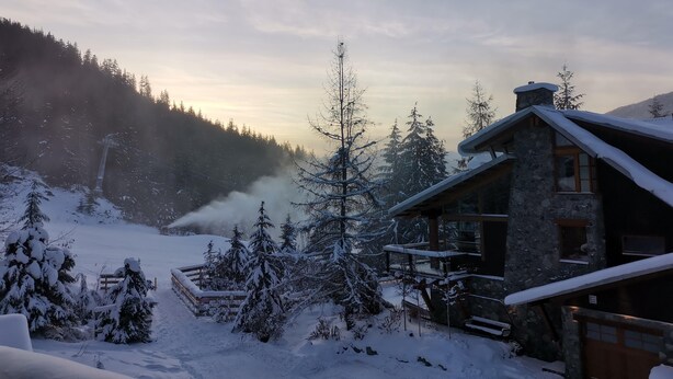 A stone cabin with a snow-covered roof, surrounded by snow-laden trees and a mountainous backdrop.