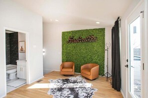 Ein modernes Wohnzimmer mit grüner Wand, zwei Ledersesseln, einem Kuhfellteppich und einem durchgegangenen Tür erkennbaren Badezimmer.