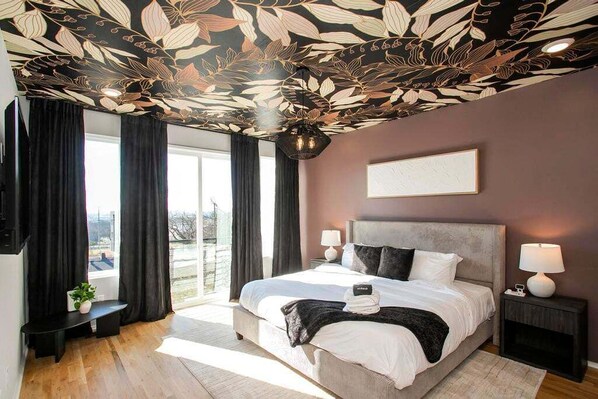 Ein modernes Schlafzimmer mit einem großen Bett, dunklen Vorhängen, einer Decke mit Blättermuster und Blick durch das Fenster.