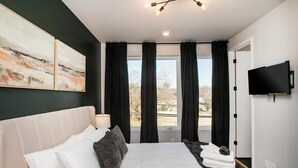 A modern bedroom with a large bed, a flat-screen TV, and a view of the outdoors through the window. - 🌇 Nashville Rooftop Retreat - Fire Pit, Skyline Views, Sleeps 12! 🎶 (Nashville)