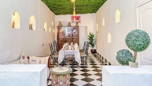 Un pasillo con piso a cuadros, un comedor con mesas y sillas, un mueble con puertas de vidrio y un techo cubierto de follaje verde.