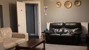 A living room with a white leather sofa, a black leather couch, and a wooden coffee table. There is a door leading to another room, a wall-mounted light fixture, and three decorative plates on the wall. - THE RATHMORE (Cambridge)