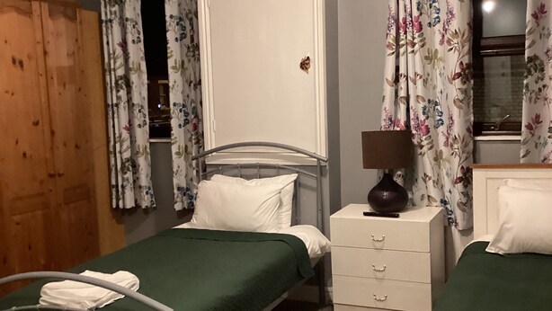 A bedroom with a single bed, a bedside table with a lamp, a wardrobe, and a window with floral curtains. - THE RATHMORE (Cambridge)