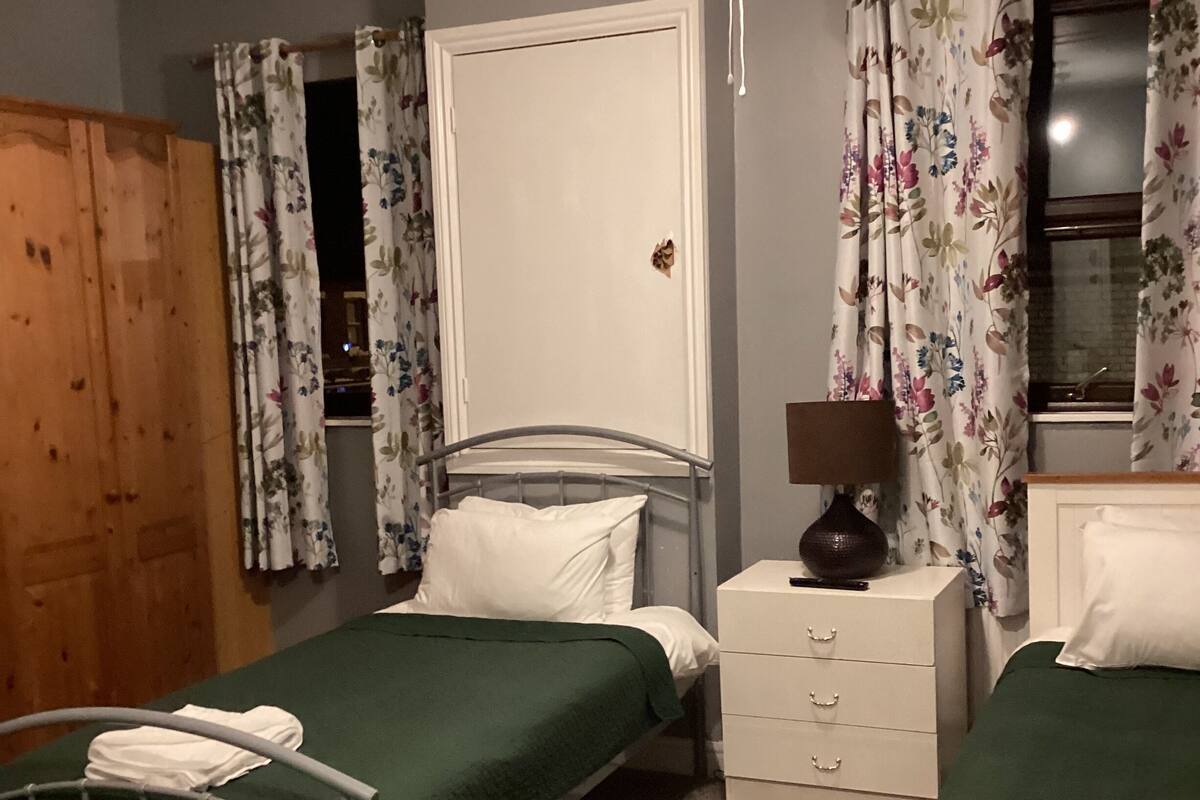 A bedroom with a single bed, a bedside table with a lamp, a wardrobe, and a window with floral curtains.