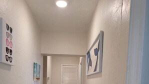 Ein Flur mit gefliestem Boden, weißen Wänden und einer Deckenleuchte. An den Wänden befinden sich Bilder in Rahmen und ein Lüftungsgitter.