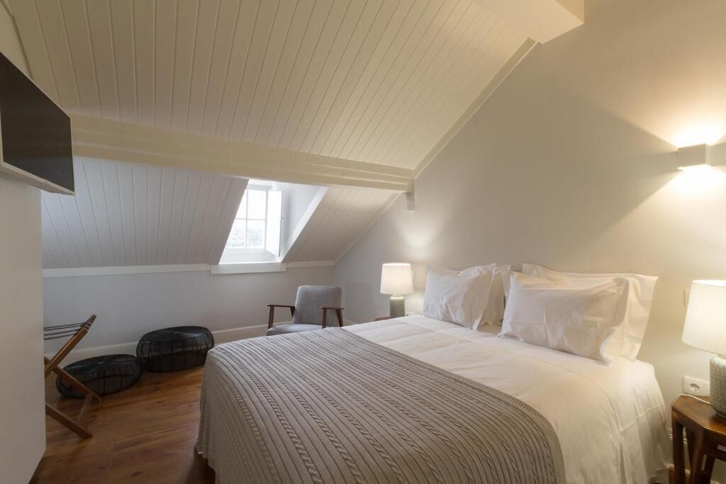 A bedroom with a sloped ceiling, a large bed with white bedding, two bedside lamps, a chair, and a window.