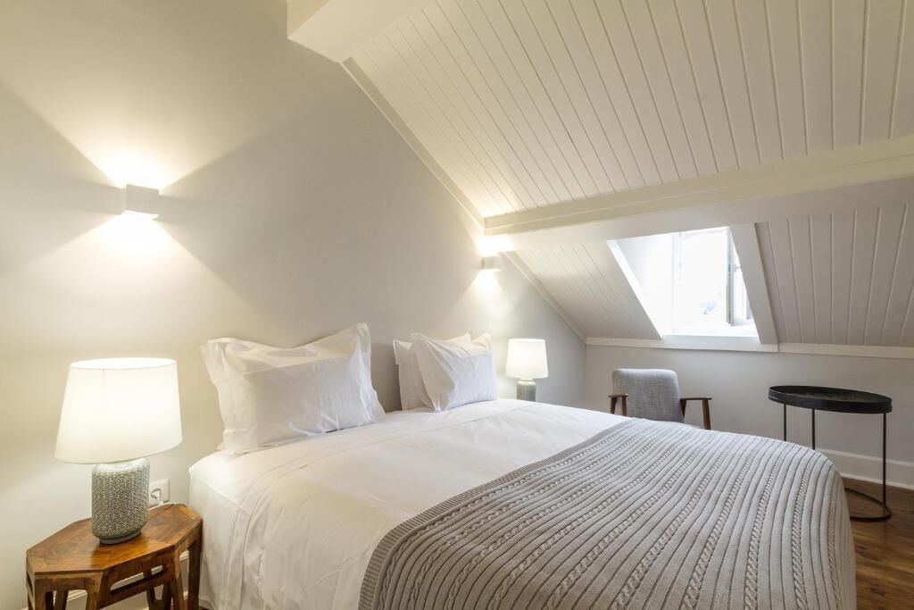 A bedroom with a sloped ceiling, a bed with white linens, a bedside lamp, and a window.