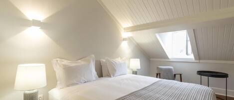 A bedroom with a sloped ceiling, a bed with white linens, a bedside lamp, and a window.