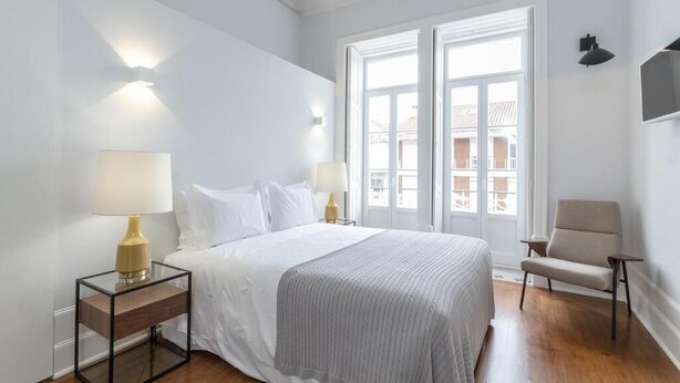Habitación de hotel con cama, dos mesitas de noche con lámparas, una silla y un ventanal con vistas a edificios.