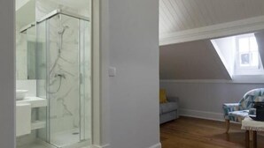 Un amplio dormitorio en el ático con tragaluces, ducha con cerramiento de vidrio, piso de madera y una zona de estar acogedora.