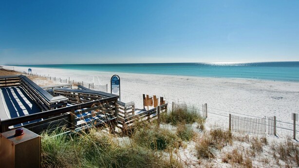 Ein Strand mit einer Holzterrasse und Liegestühlen, feiner Sand und ein klarer, blauer Himmel.