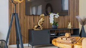 Una sala de estar moderna con un televisor de pantalla plana grande montado en una pared de madera, una lámpara de trípode, una estatua dorada, una mesa de centro de madera y un sillón de cuero.