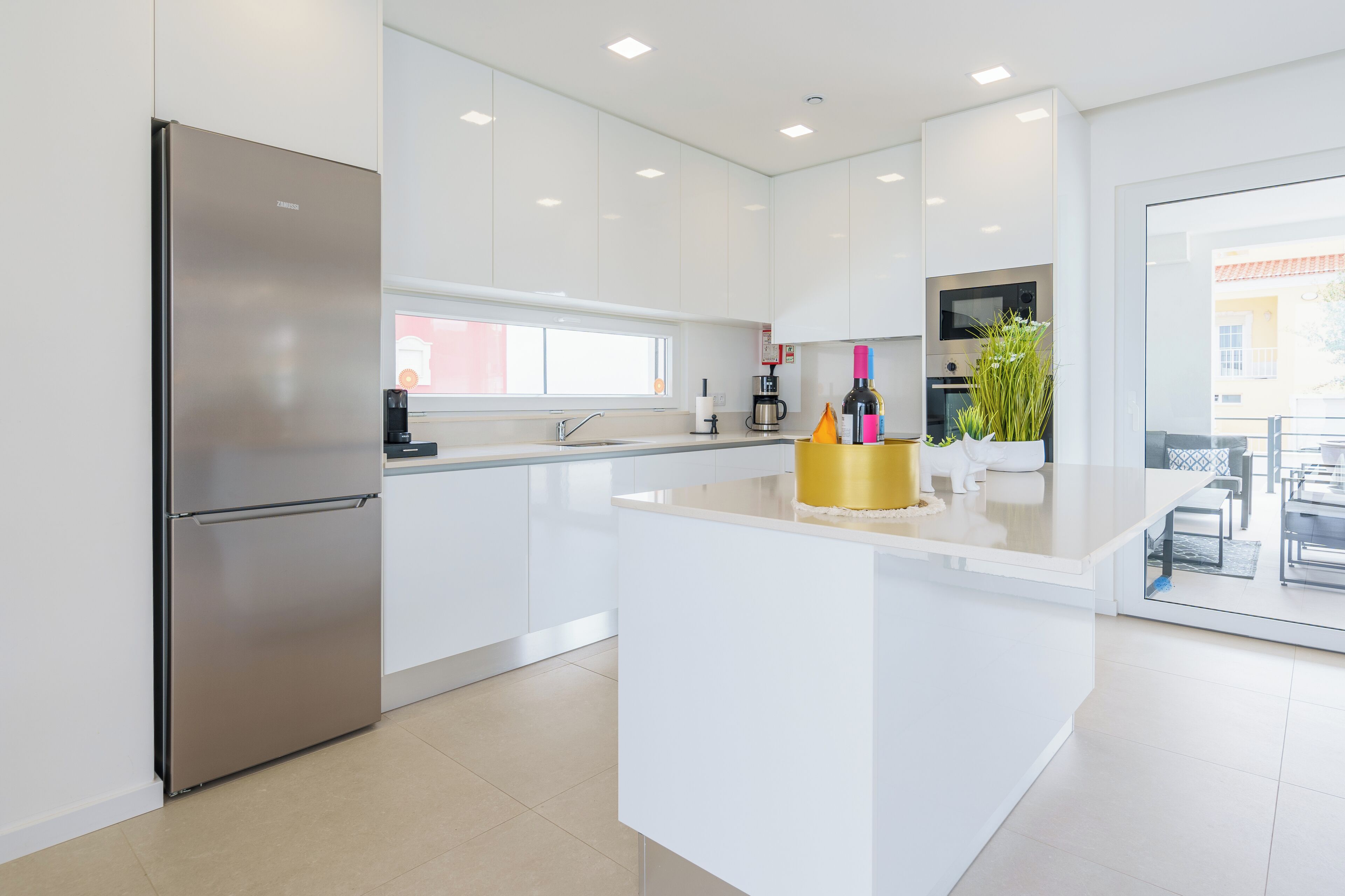 A modern kitchen with white cabinets, a stainless steel refrigerator, a central island, and a view of the outside.